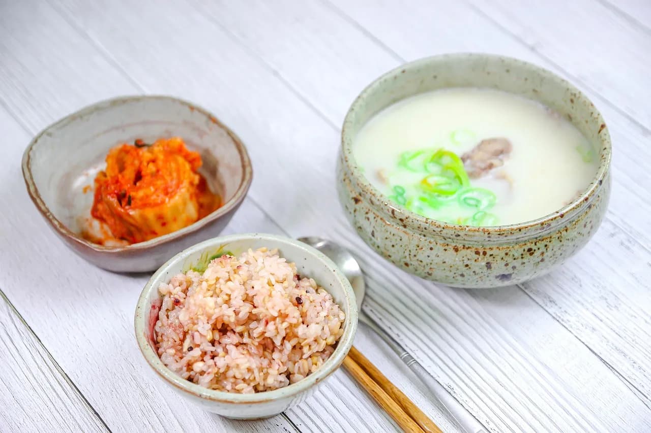 A bowl of milky white broth with sliced green onion, served with kimchi and a bowl of mixed rice