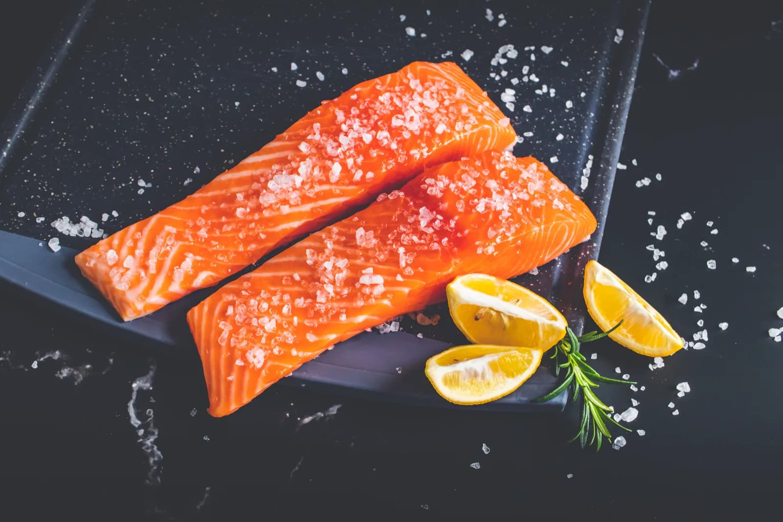 Two raw salmon fillets with coarse salt, lemon wedges and a sprig of rosemary on a dark slate