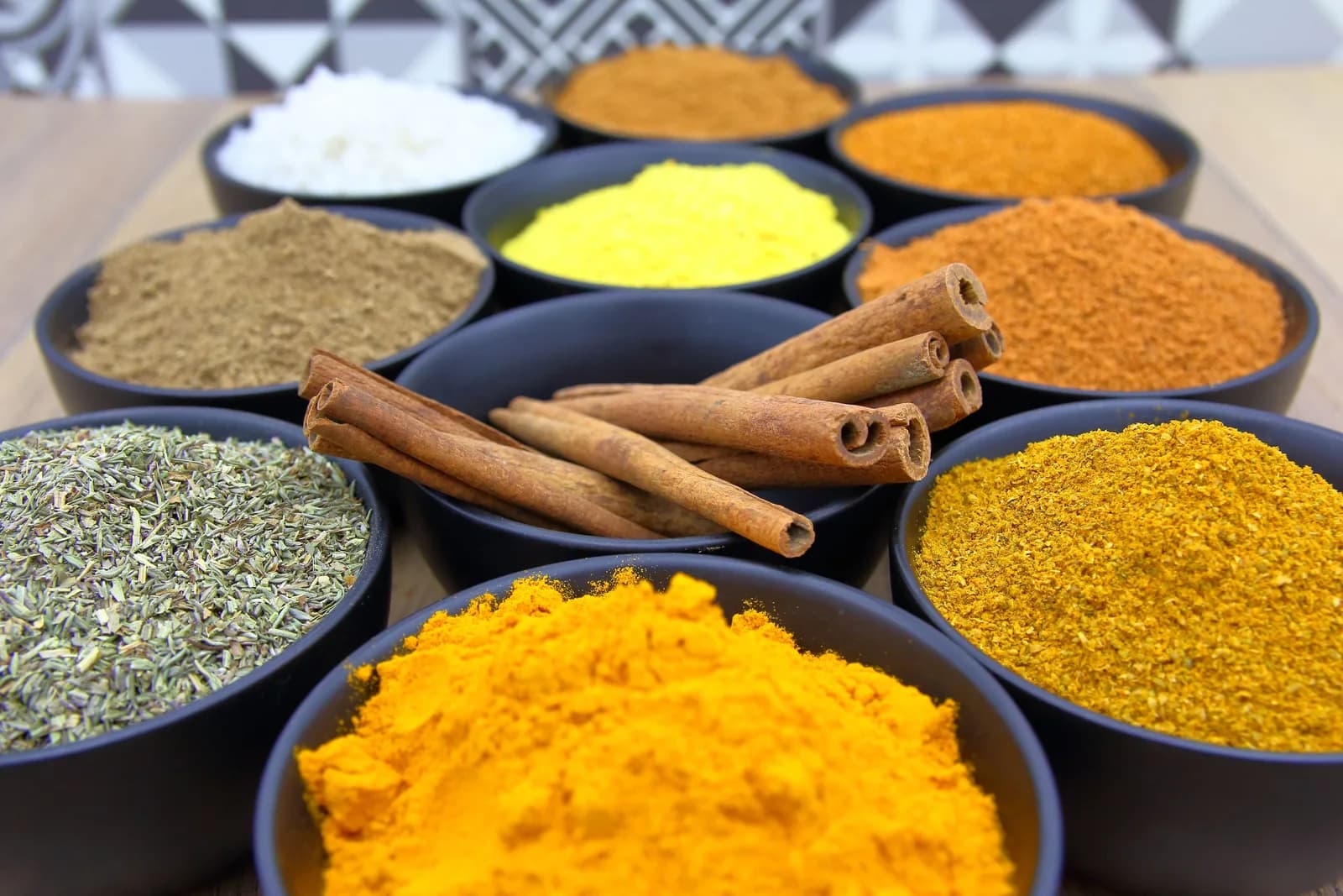 Black bowls of ground spices in yellow, orange, green and brown, with cinnamon sticks laid across the center