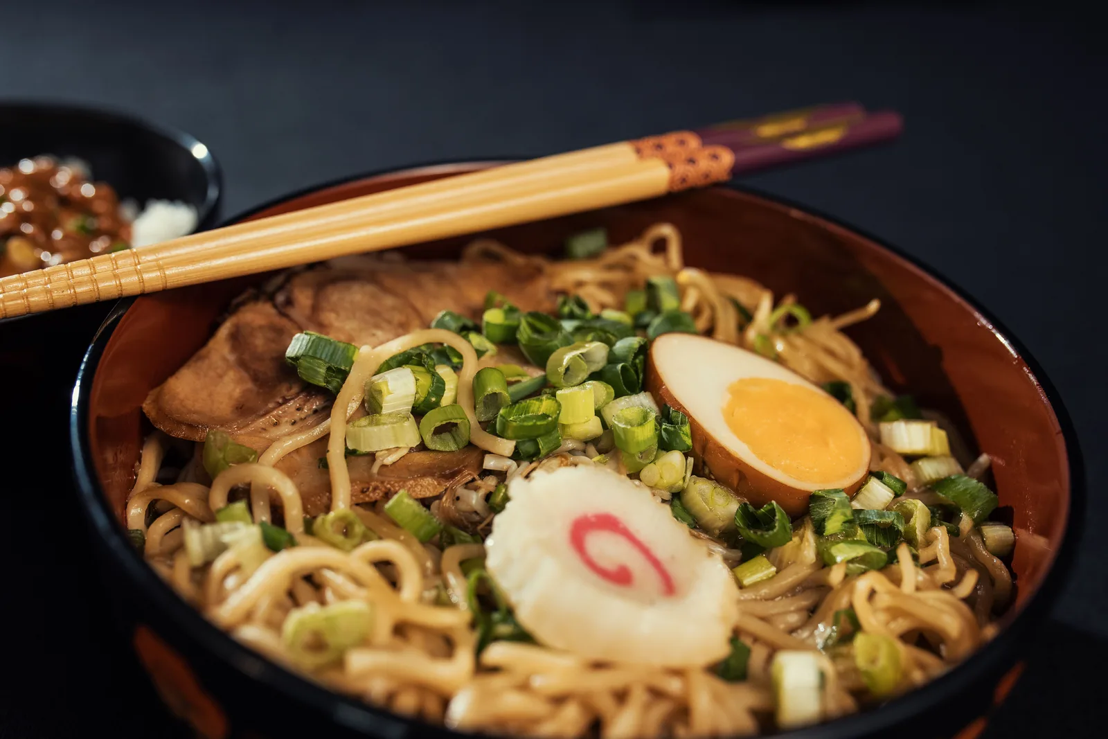 A bowl of ramen with wavy yellow noodles, sliced spring onion, a soft boiled egg and a slice of narutomaki