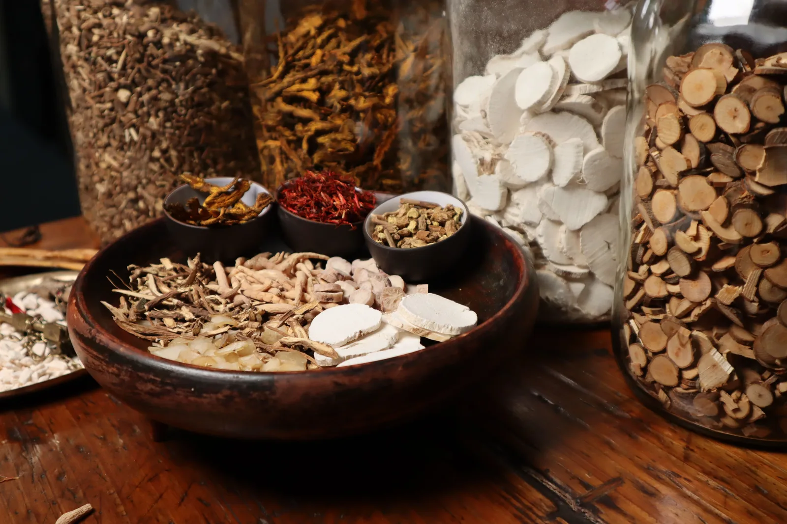 Dried Chinese medicinal roots, bark and herbs in a wooden bowl, with more in tall glass jars behind