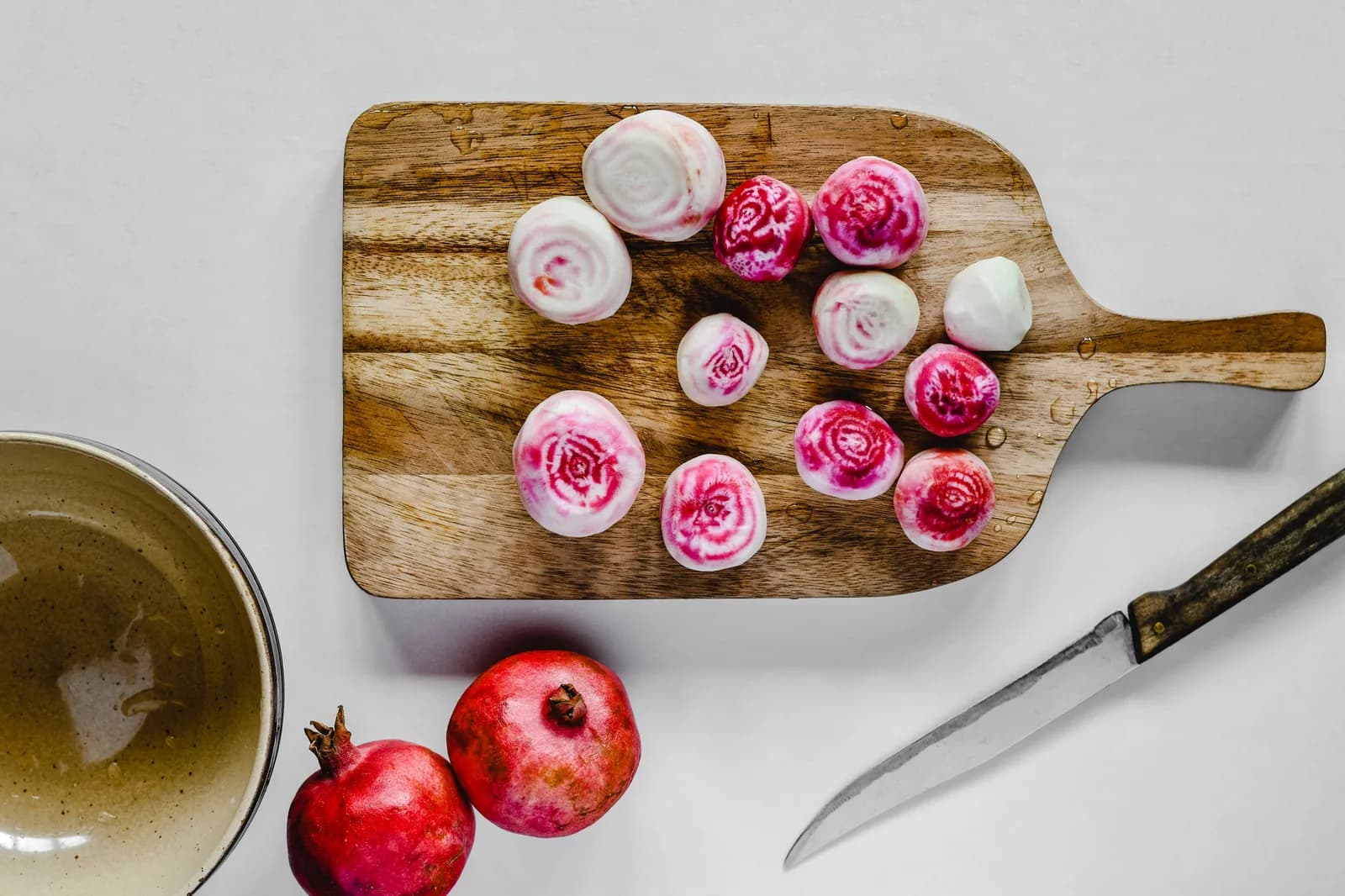 Sliced Chioggia beets on a wooden board beside two whole pomegranates and a knife