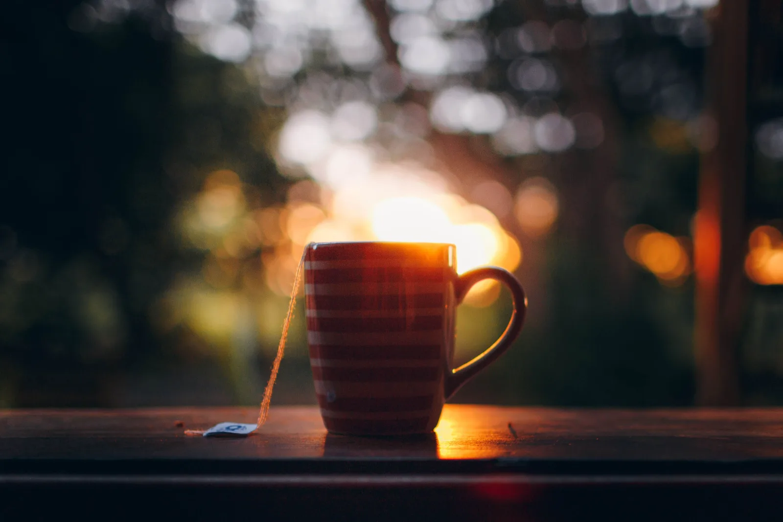 A steaming mug of tea on a wooden windowsill at sundown, the tea bag tag hanging over the side