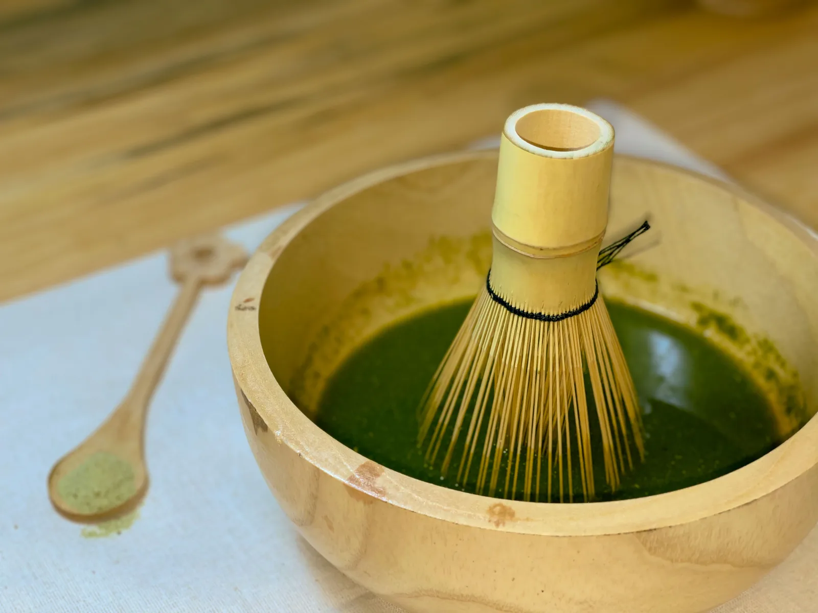 A bamboo whisk resting in a wooden bowl of frothy matcha, a spoon of matcha powder beside it