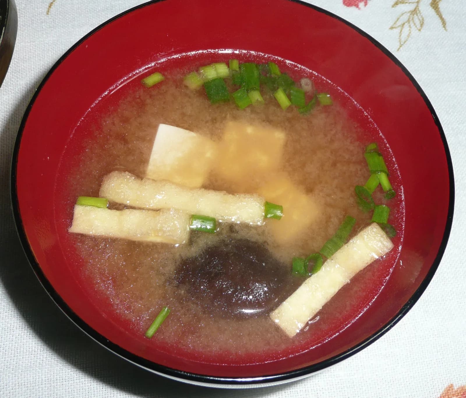 Miso soup in a red bowl with tofu, strips of fried tofu, a shiitake mushroom and sliced green onion