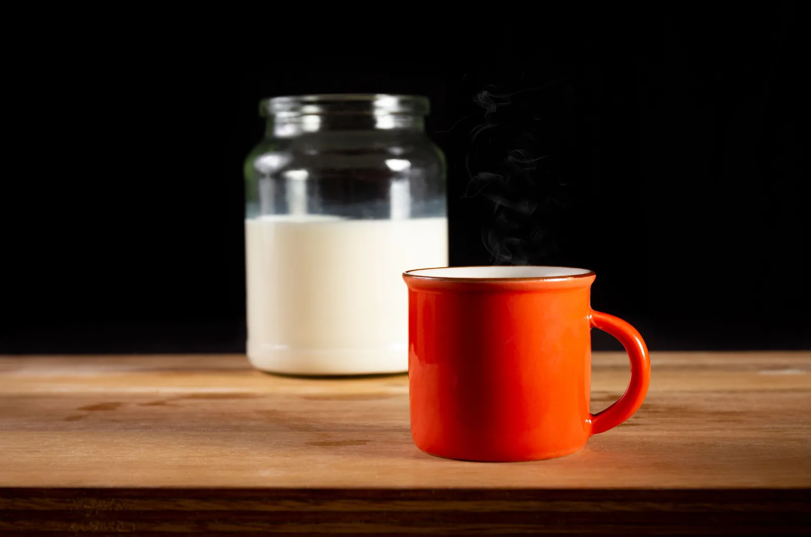 A red enamel mug beside a glass jar of milk on a wooden table, against a dark background