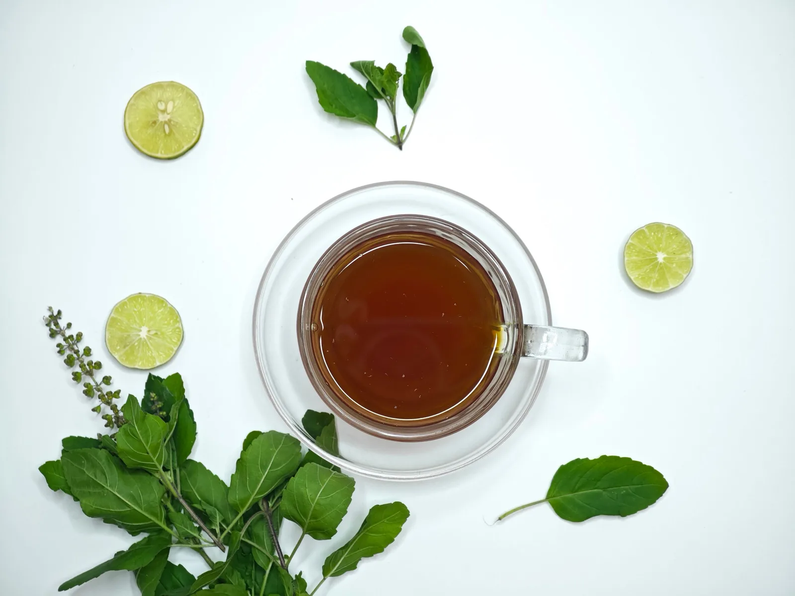 A glass cup of amber herbal tea on a white table, surrounded by sprigs of holy basil and lime slices