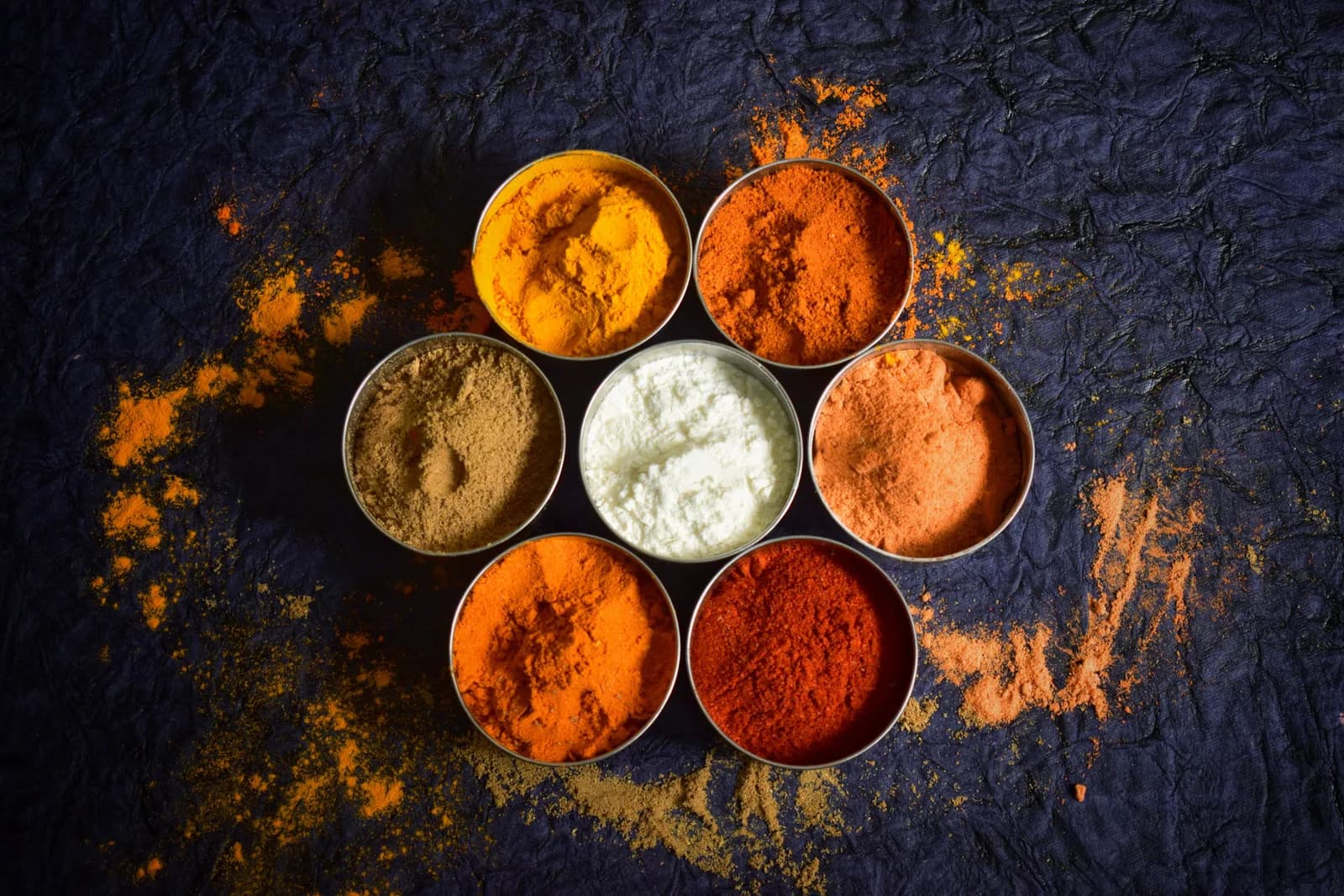 Seven small tins of ground spices arranged in a circle on dark slate, dusted with turmeric