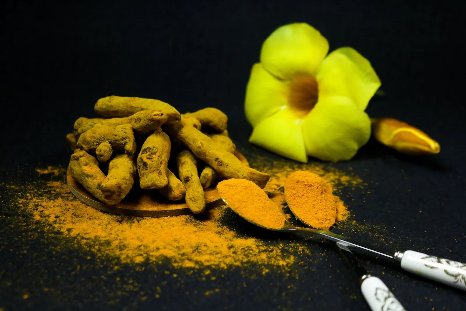 Dried turmeric roots and turmeric powder on a dark table, with a yellow flower beside them