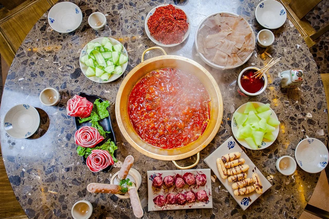 Overhead view of a hot pot table: a pot of red chilli broth in the middle, surrounded by plates of sliced meat, cucumber and small sauce bowls