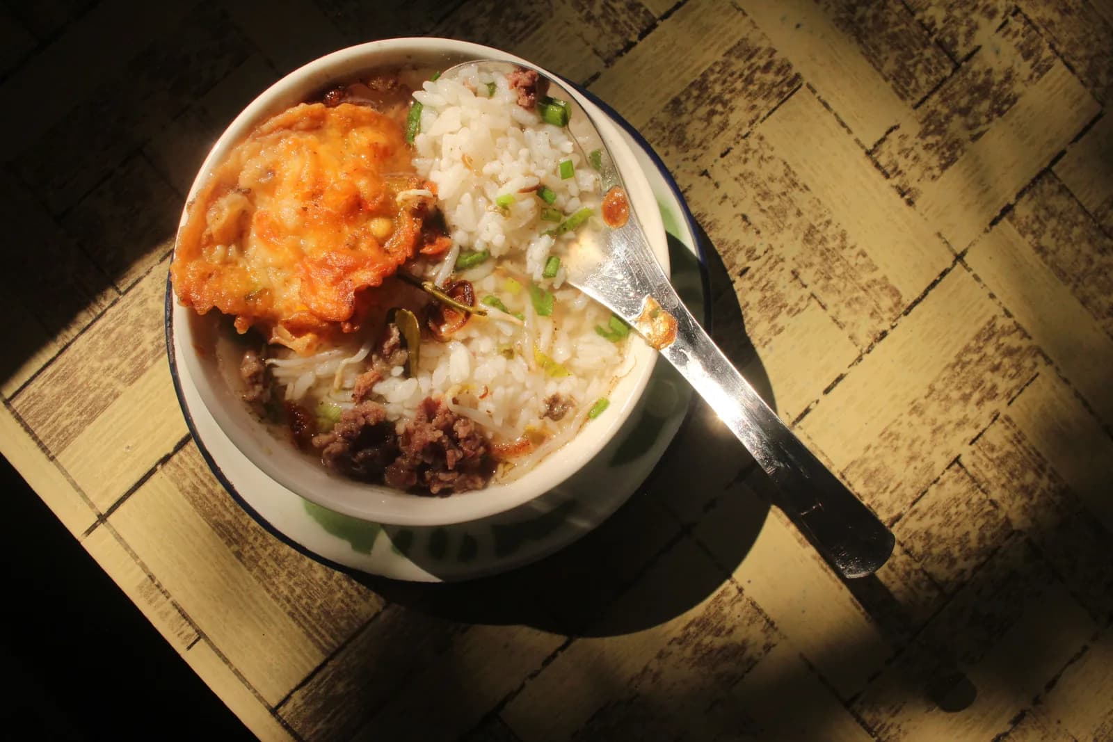 A bowl of soft rice porridge with a golden fried topping, minced meat and green onion, a spoon resting in it
