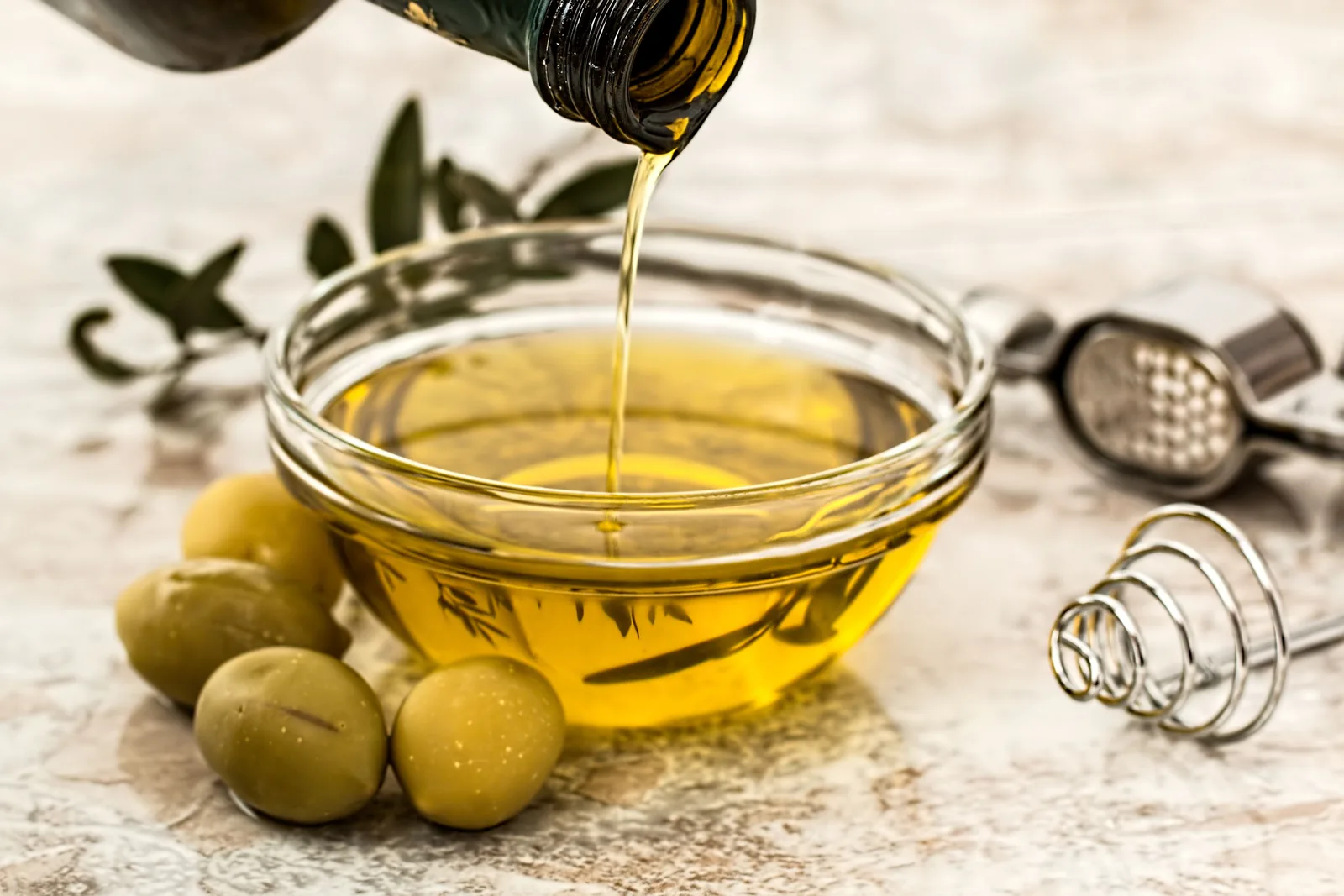 Olive oil pouring from a bottle into a glass bowl, with green olives and a garlic press beside it
