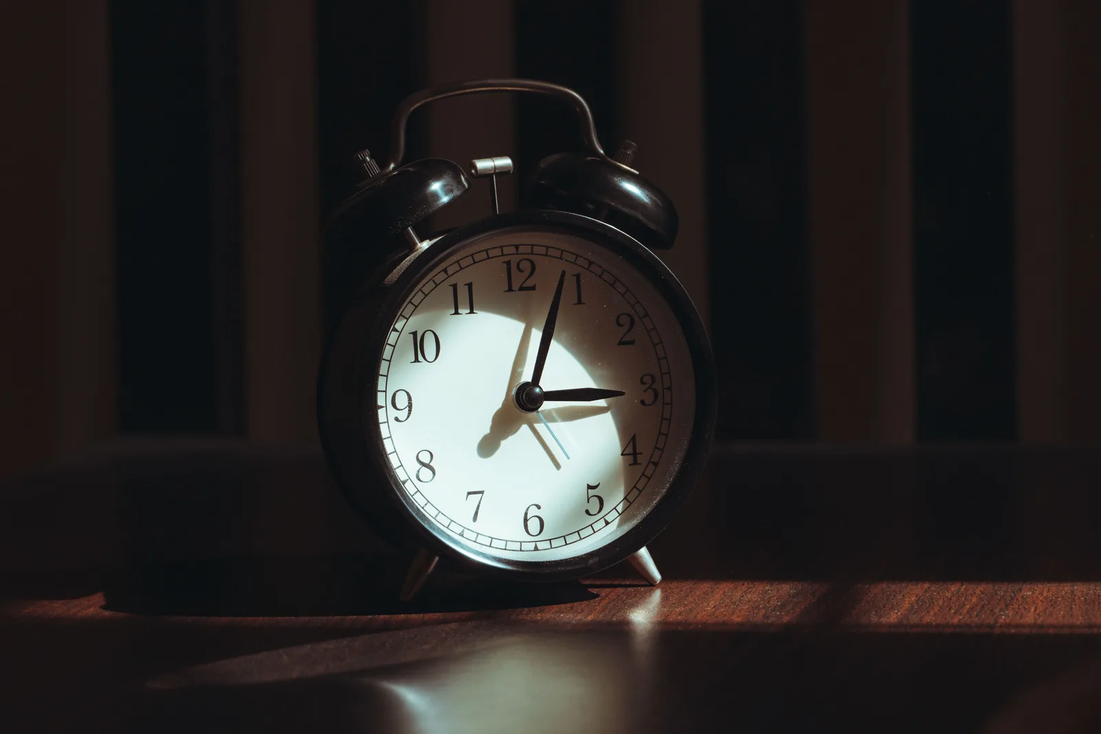 A black alarm clock on a dark wooden table in a dim room, its hands at about three o'clock