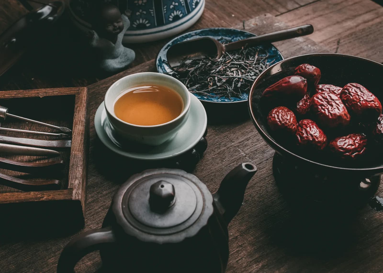 Dried red dates in a dark bowl beside a cup of tea, loose tea leaves and a clay teapot
