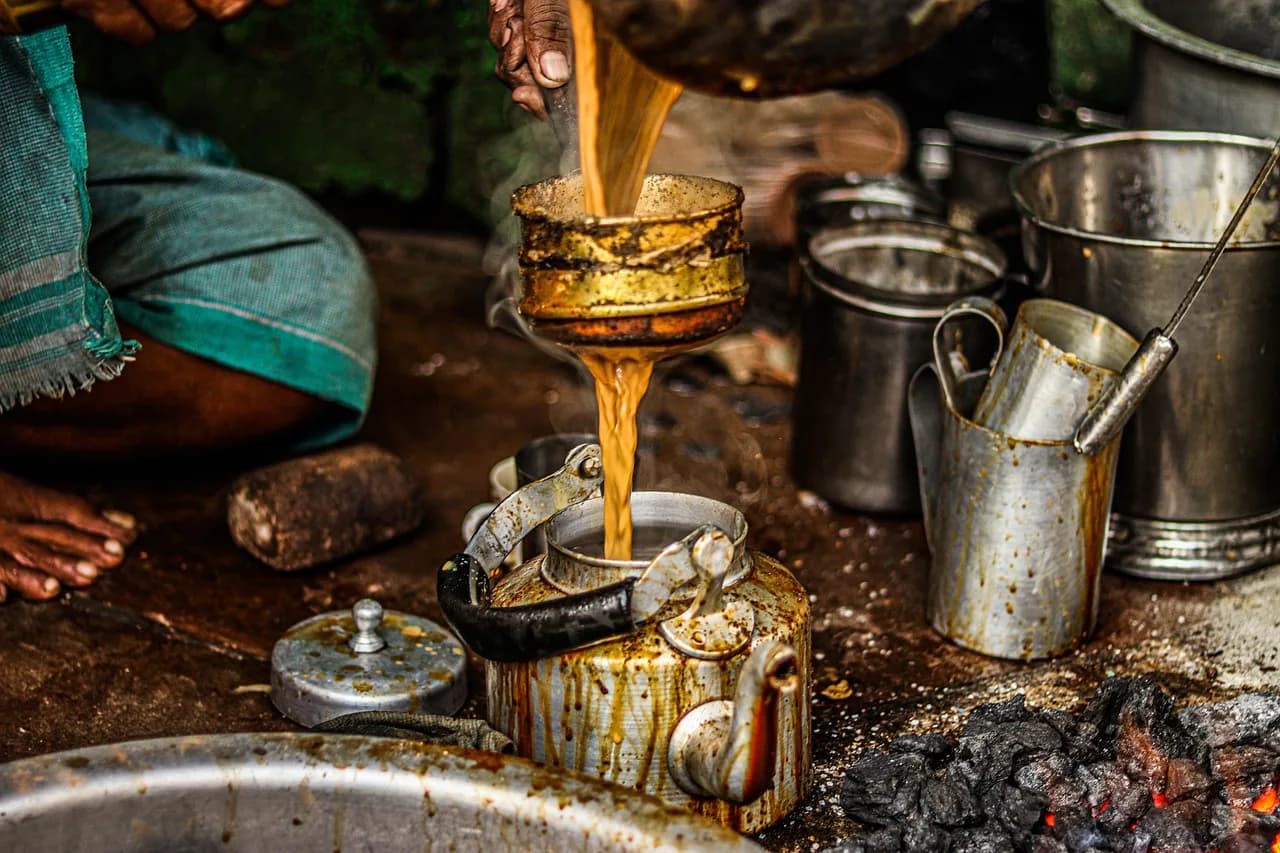 Chai being poured through a strainer into a kettle at a street tea stall, steel pots crowded around a charcoal stove