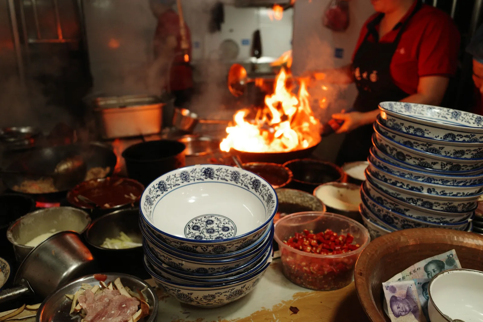 Flames leap from a wok in a Chinese kitchen, beside stacks of blue and white bowls and a dish of chopped red chillies
