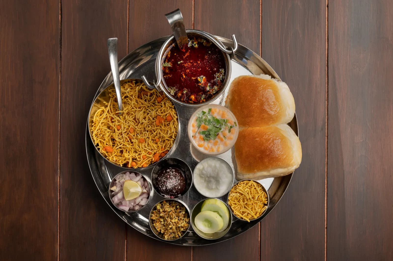 A steel thali on a wooden table with spiced curry, crunchy farsan, pav rolls, chopped onion and lemon in small bowls