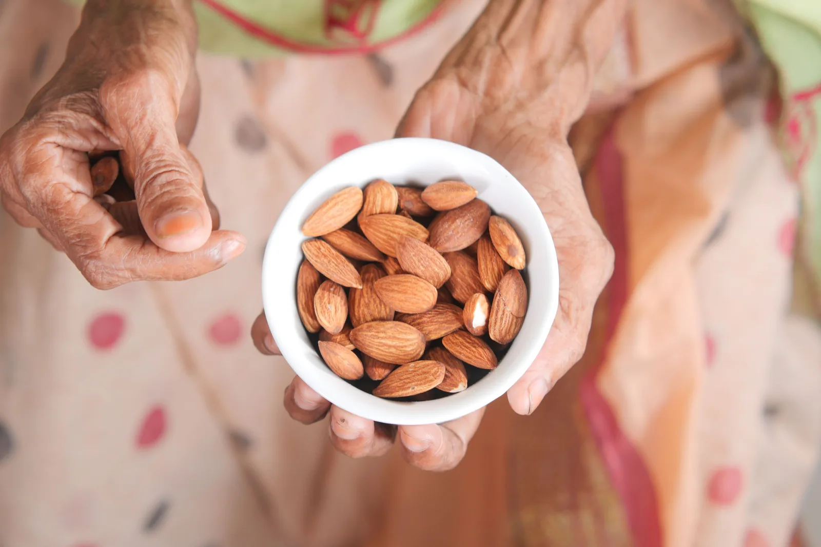 An elderly woman's hands holding a small bowl of almonds