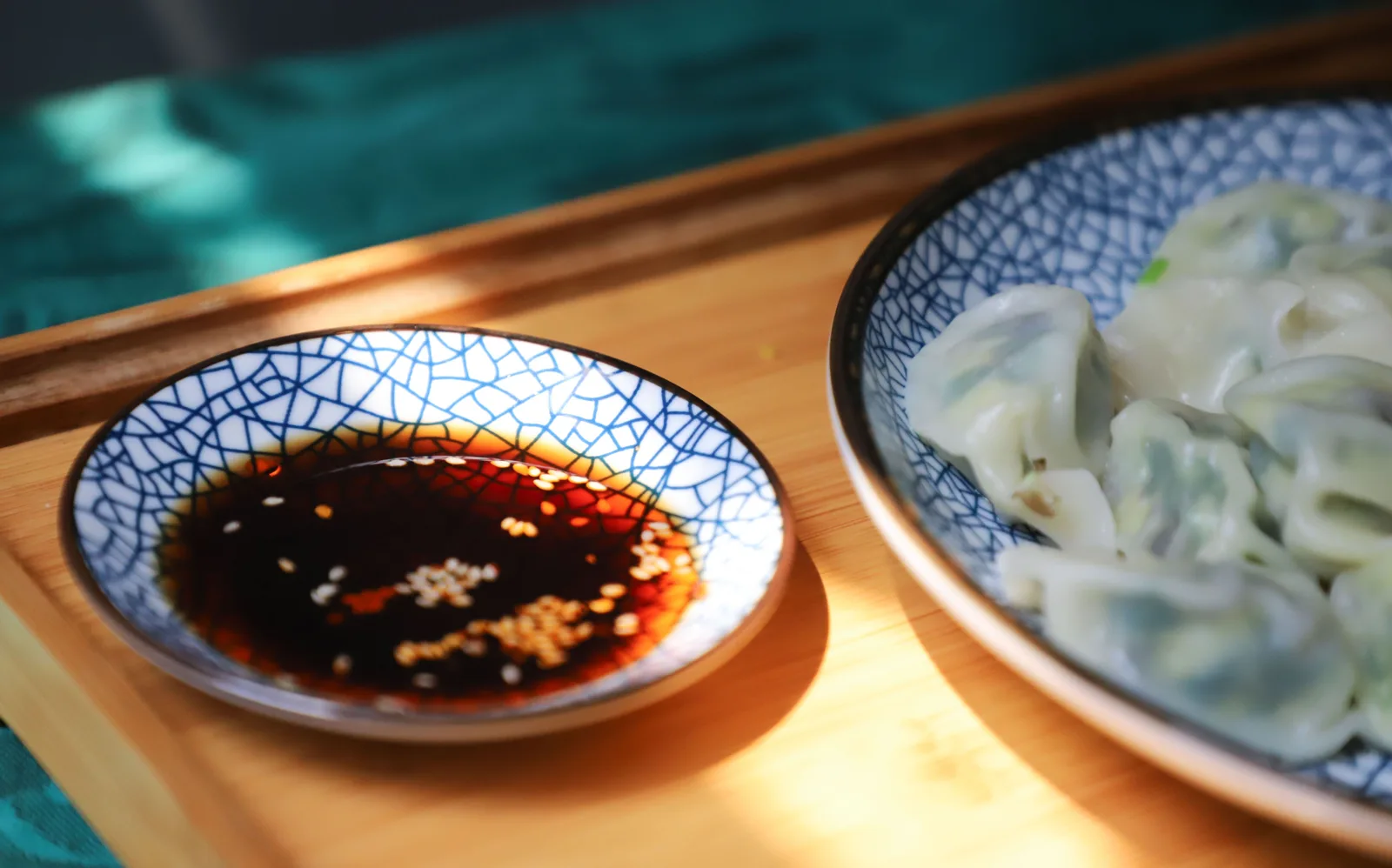 A patterned saucer of soy sauce with sesame seeds, next to a plate of dumplings on a wooden tray