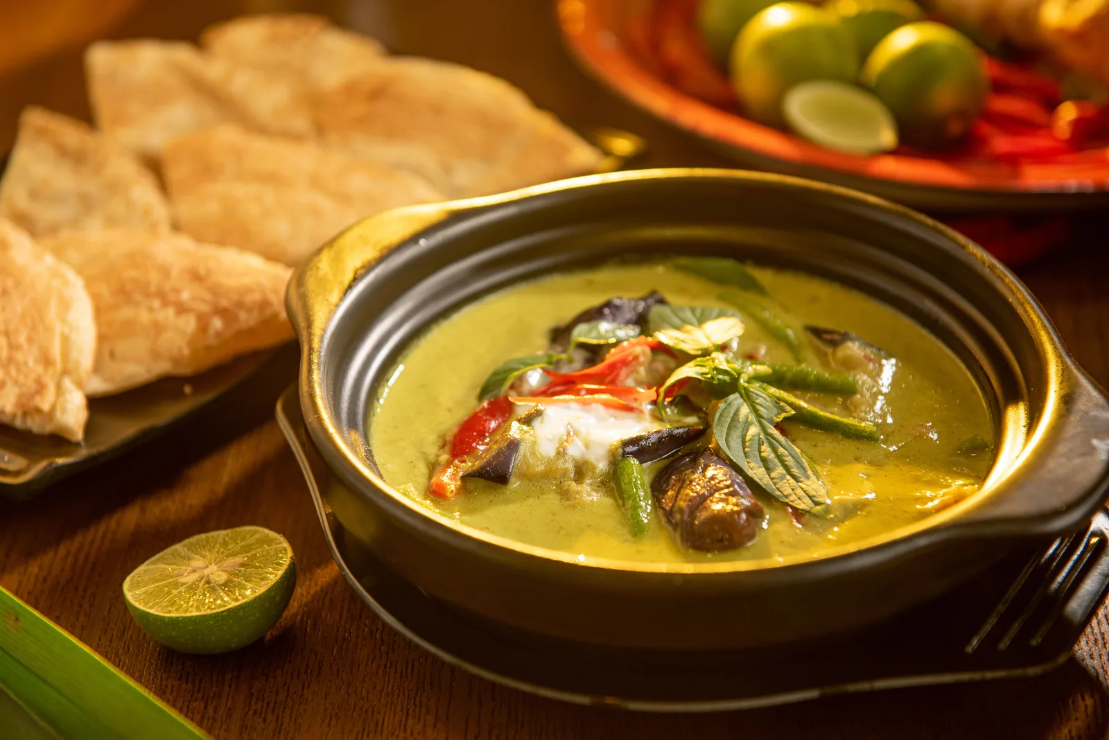 Thai green curry with eggplant and basil in a clay pot, with roti and lime on the side