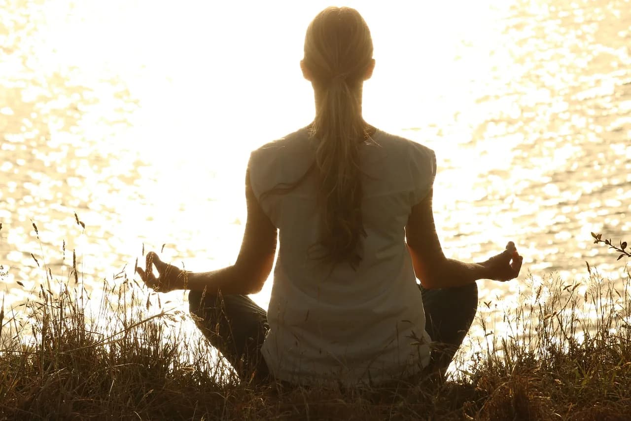 A woman seen from behind, sitting cross-legged in meditation facing water lit by low, bright sun