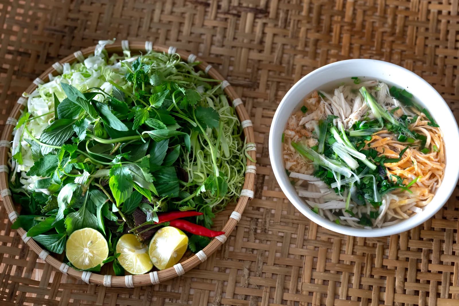 A woven tray piled with fresh herbs, lettuce, lime halves and red chillies next to a bowl of noodle soup
