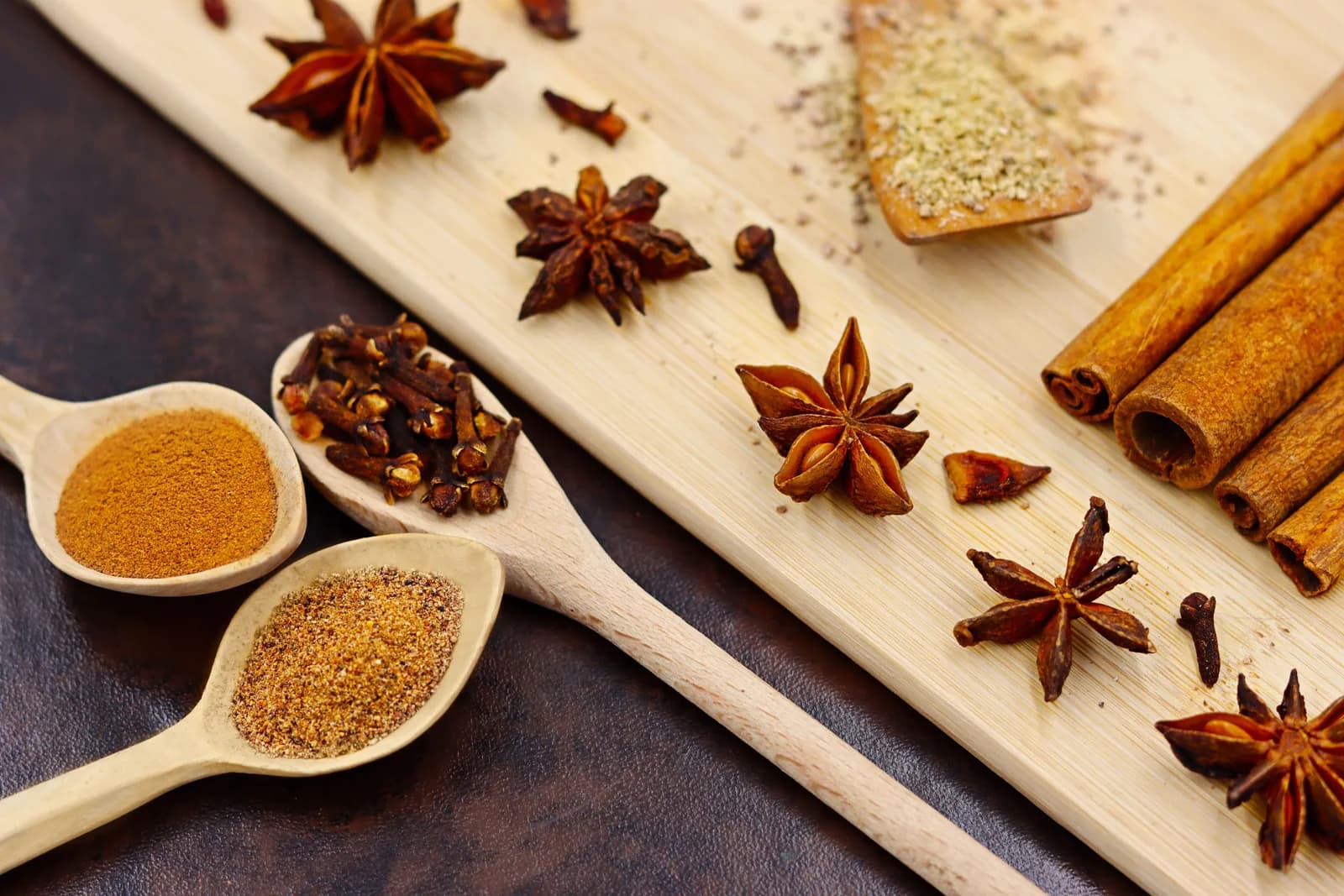 Star anise, cloves and cinnamon sticks on a wooden board, with ground spices in two spoons