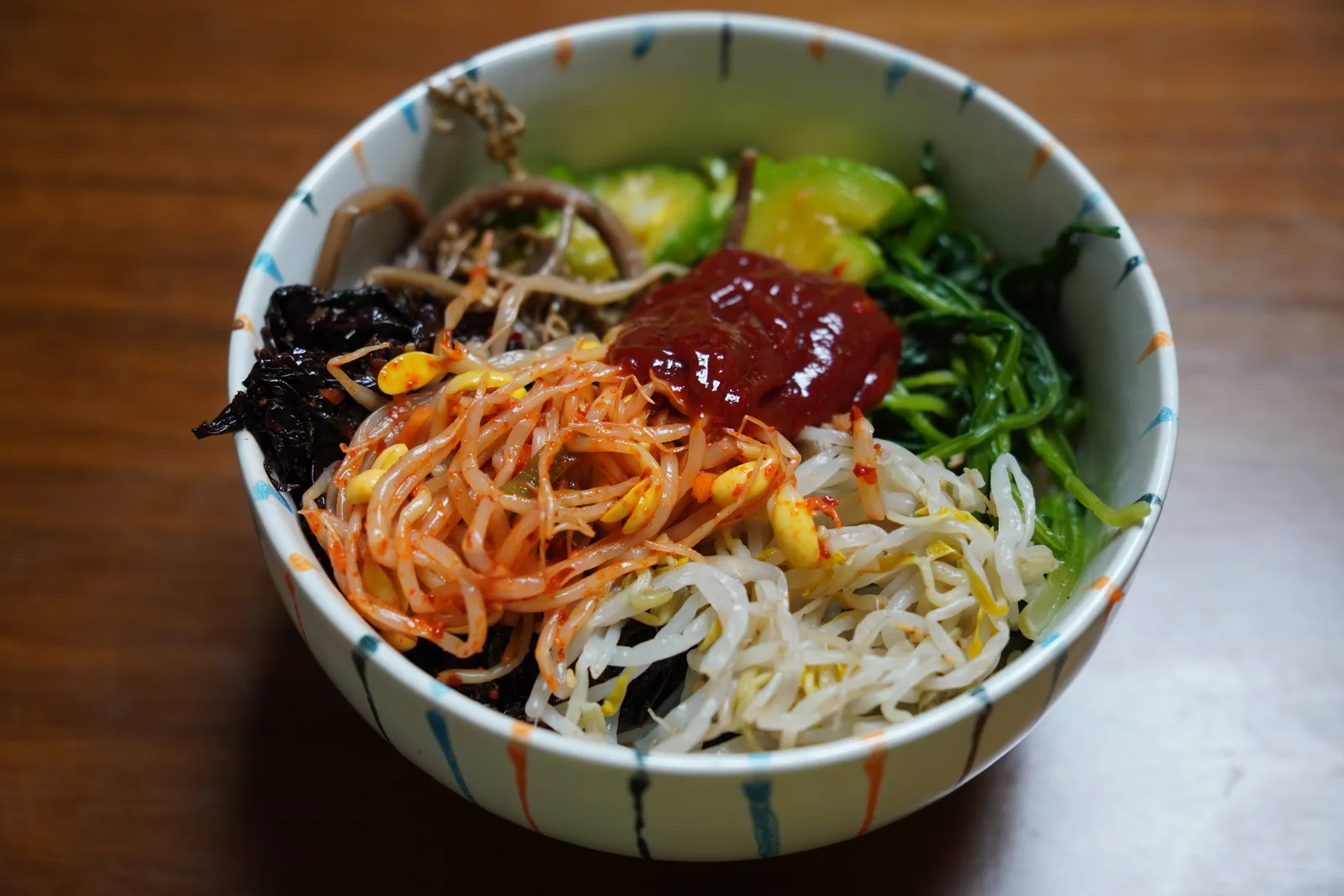 A bowl of bibimbap topped with a spoonful of red gochujang over bean sprouts, carrots, greens and seaweed