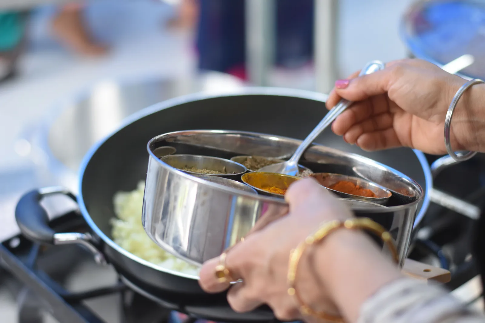 A cook spoons spice from a round steel spice box into a pan on the stove