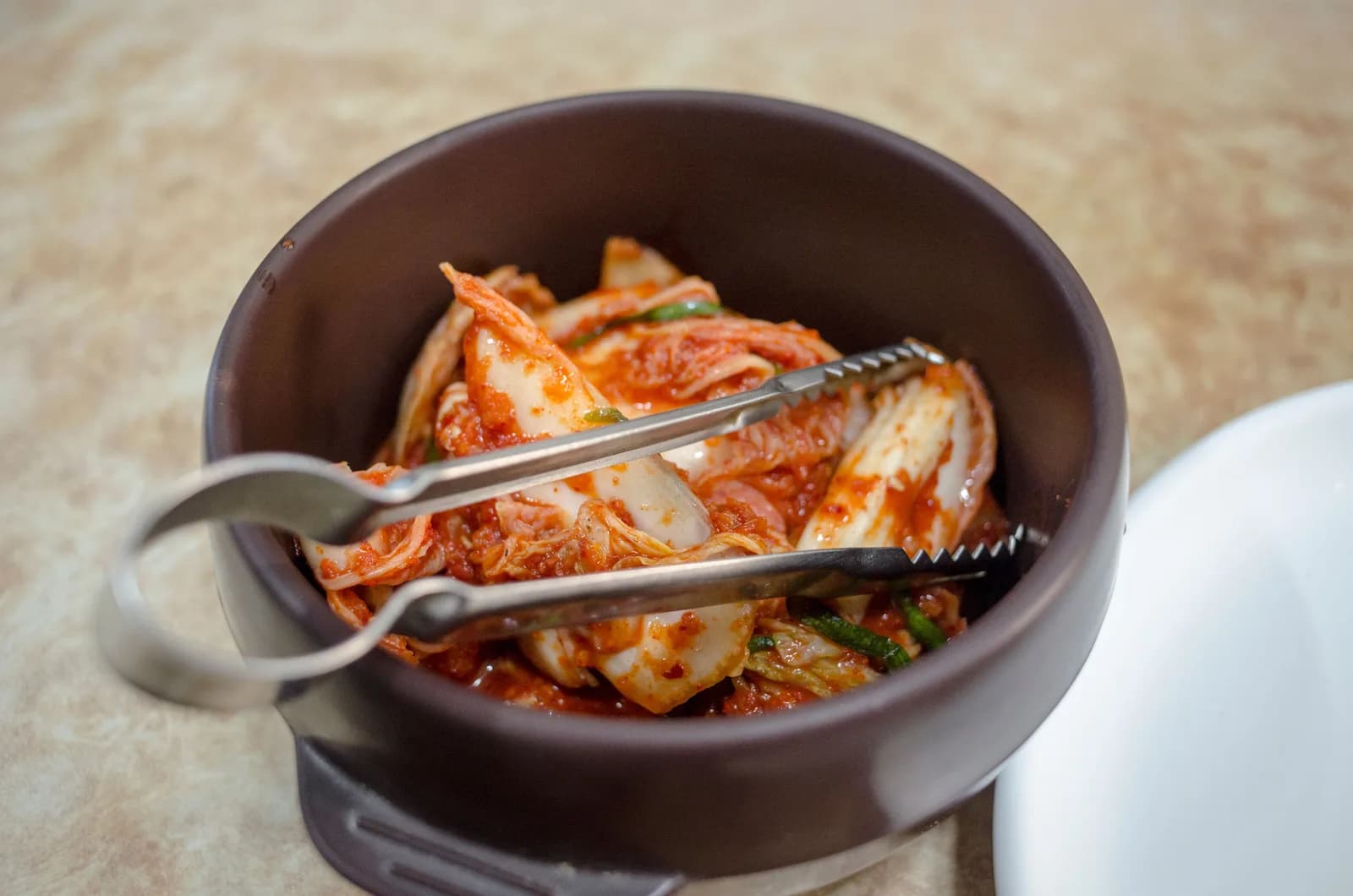 Napa cabbage kimchi in a dark ceramic bowl with metal serving tongs
