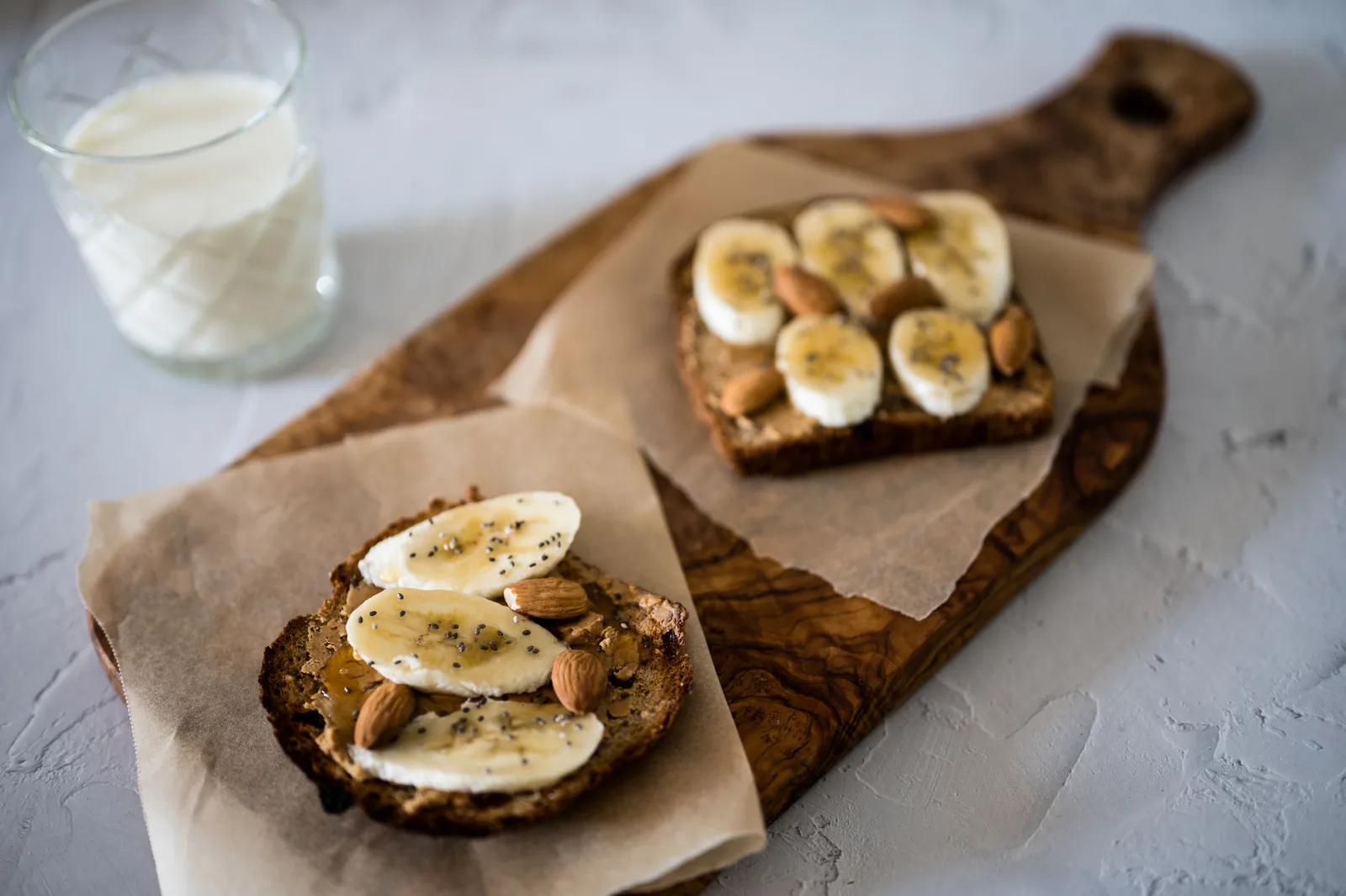 Two slices of toast spread with nut butter and topped with banana and almonds, with a glass of milk behind them