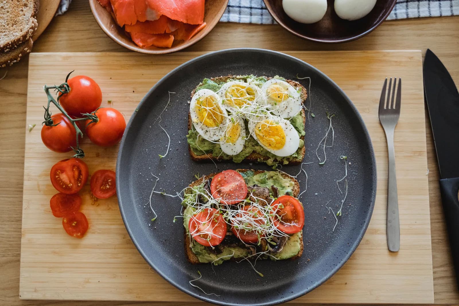 Toast topped with avocado, sliced boiled egg and tomato, with smoked salmon and cherry tomatoes on a wooden board