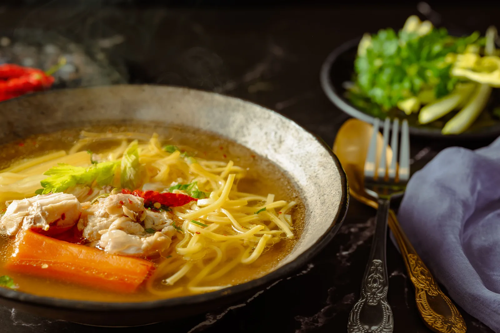A bowl of chicken noodle soup with carrot and red chilli, a dish of chopped green onions behind it