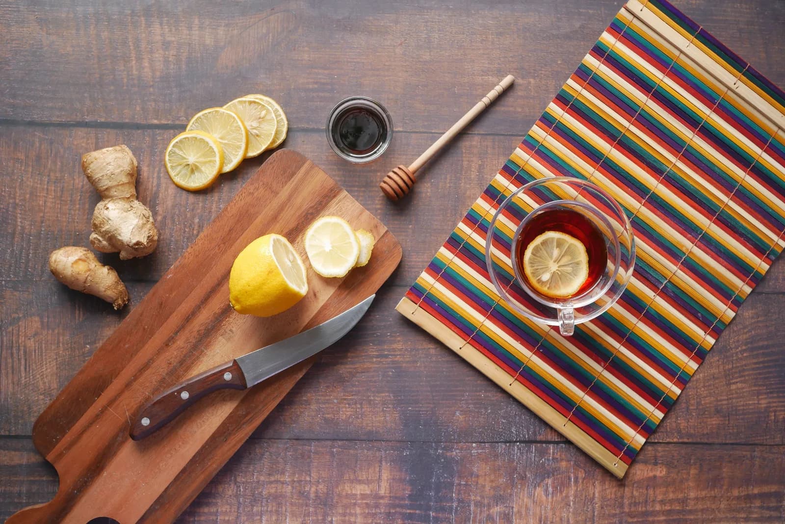 A glass of hot tea with a lemon slice, next to fresh ginger, sliced lemon and a honey dipper on a cutting board