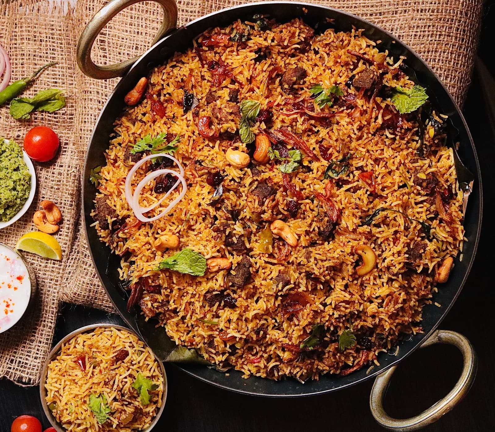 A wide pan of biryani with long, separate grains of rice, onion rings, cashews and mint, seen from above