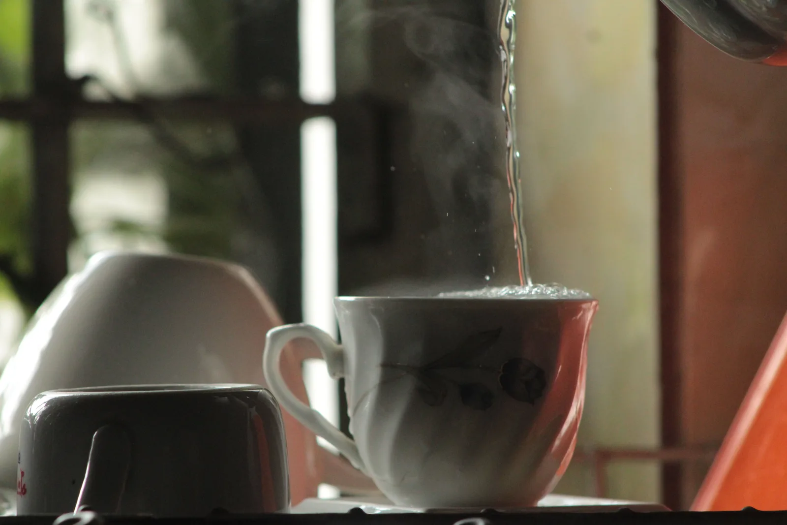 Hot water poured into a white teacup, steam rising by a window