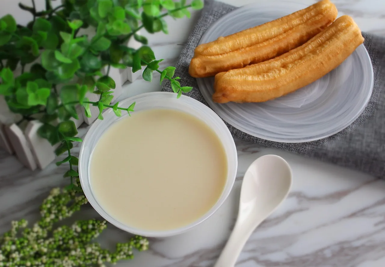 A bowl of warm soy milk with a spoon beside two fried dough sticks on a grey plate