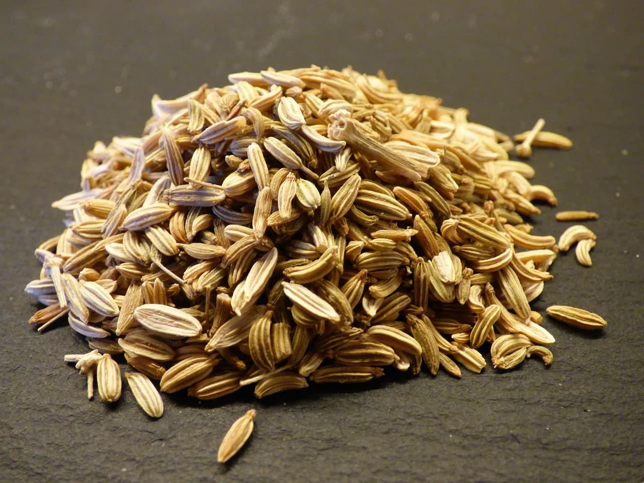 A small pile of dried fennel seeds on a dark slate surface