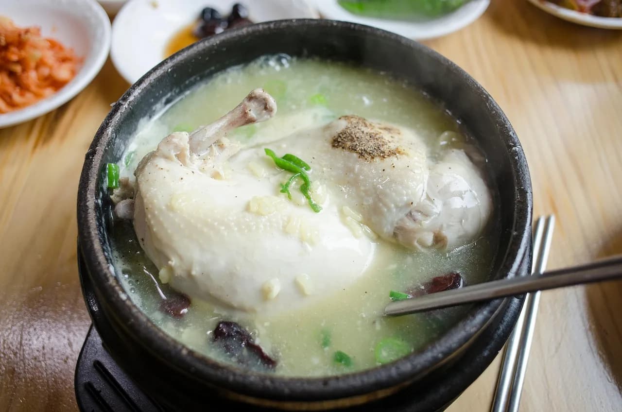 Samgyetang in a black stone bowl, a whole young chicken in milky broth with jujube and scallion, banchan behind