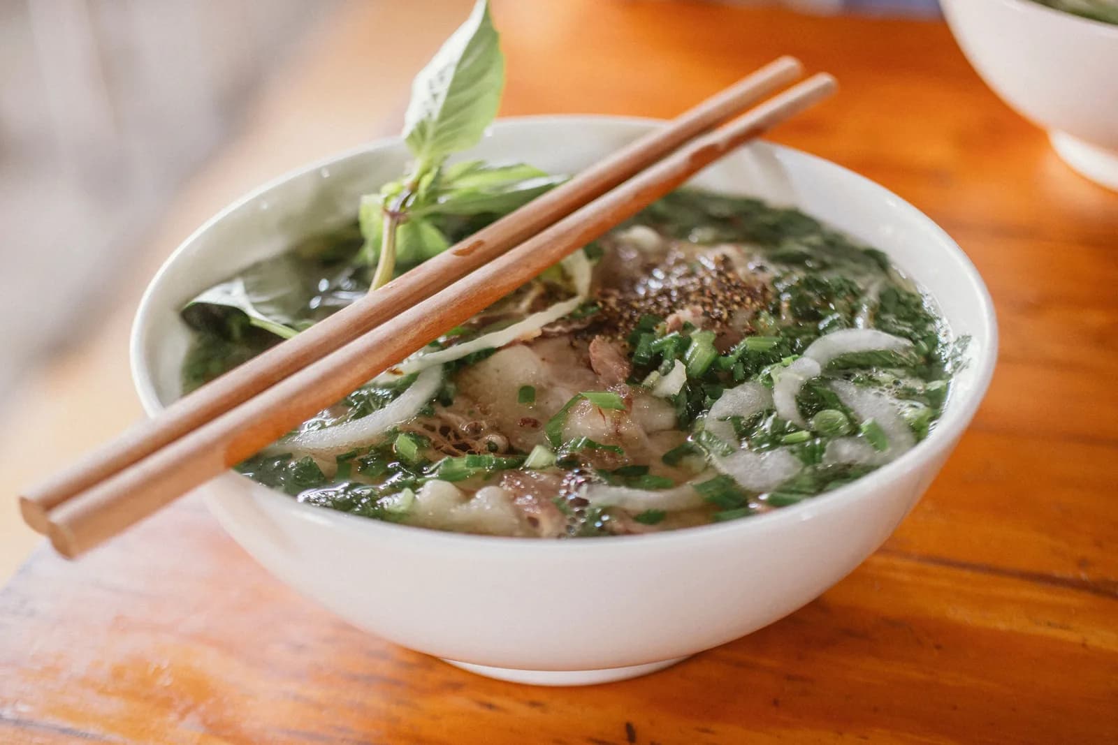 A bowl of pho topped with herbs and onion, with wooden chopsticks resting across the rim